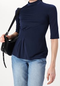 Navy blue fitted top with a high neckline, short sleeves, and gathered detailing at the waist, paired with light blue jeans and a black handbag.