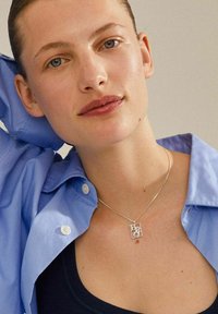 Silverhalsband med geometrisk design, synlig kedja, bärs över en mörk topp, i kombination med en ljusblå skjorta med knappar.