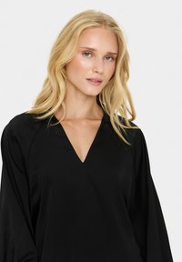 Black blouse with a V-neck, long sleeves, and a smooth, textured fabric. Features slight draping detail at the shoulders for an elegant silhouette.