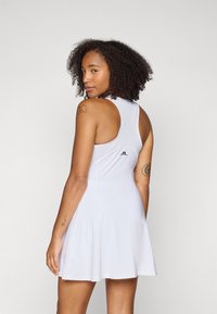 Moteris su garbanotais plaukais, vilkinti balta be rankovių suknelę su tinkline nugarėle, demonstruojanti kelias rankų tatuiruotes, žiūrinti per petį.