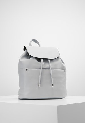 Sac à dos en cuir gris clair avec fermeture à rabat, cordon de serrage, et accents en métal argenté. Texture lisse, design minimaliste, taille moyenne.