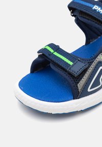 Sandales bleu marine avec des brides en tissu, dotées d'une bande d'accent vert vif. Semelle intérieure bleue texturée et semelle en caoutchouc blanche.