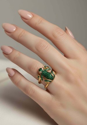 Hand with nude polished nails wearing a gold ring shaped like a frog encrusted with small green gemstones on the middle finger.