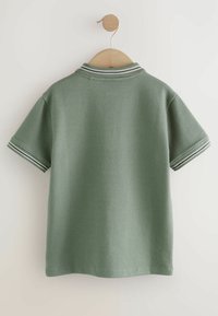 Polo verde con tessutoTexture, maniche corte e colletto a righe. Appeso a una gruccia di legno su uno sfondo chiaro.