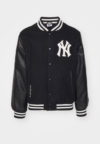 Svart varsityjacka med läderärmar, vitrandig krage och manschetter, tryckknappar och vit New York Yankees-logga på bröstet.