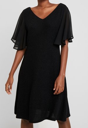 Robe noire mi-longue avec manches transparentes flottantes et col en V portée par une personne debout sur un fond gris clair.