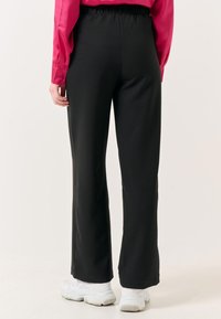 Zwarte, hoge broeken met een relaxed fit, voorzien van een elastische tailleband. Gecombineerd met een roze shirt met lange mouwen en witte sneakers.