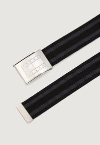 Čierny tkaný opasok so sivými pruhmi, vybavený štvorecovou striebornou kovovou sponou s gravírovaným nápisom "TOMMY JEANS." Hladká textura a obdĺžnikové konce.