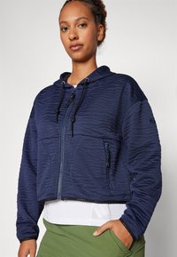 Mørkeblå, kort hoodie med teksturert stoff, glidelås foran, justerbar hette med snøring og en lomme som detalj. Laget over en hvit topp.