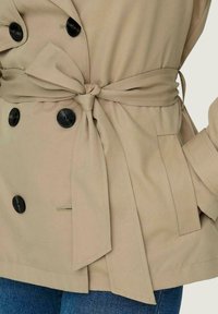 Trench coat beige doppiopetto con cintura legata in vita, bottoni neri e tasche laterali; presenta un tessuto liscio e testurizzato.