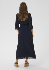 Marineblaue, plissierte Maxikleid mit langen, transparenten Ärmeln und einem gerafften Taillenbereich. Das Kleid hat einen fließenden Rockteil und wird mit Sandalen auf Korkabsatz kombiniert.