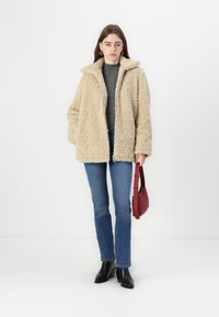 Beige bundový kožich z umělé kožešiny s klopovým límcem, kombinovaný se šedým topem a modrými džíny. Modelka drží červenou tašku a má na sobě černé boty.