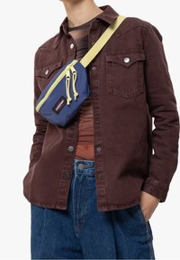 Námorně modrá crossbody taška se žlutým zipem a stahovacími šňůrkami, s logem. Nositá přes hnědou košili, stylizována s modrými džíny.