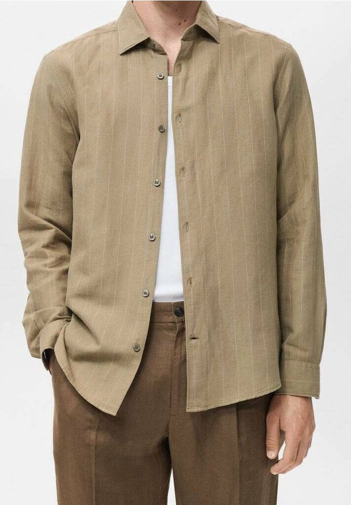 Homme portant une chemise beige à fines rayures boutonnée sur un t-shirt blanc, associée à un pantalon marron, debout avec une main dans la poche.