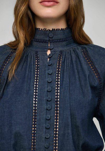Femme portant un chemisier en denim foncé à col montant avec des inserts en dentelle verticale et des boutons recouverts de tissu sur le devant et au col.