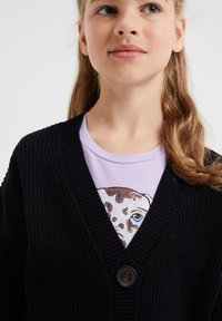 Cardigan nero a maglia con scollatura a V profonda, dotato di chiusura frontale con bottoni. Indossato sopra una t-shirt di colore viola chiaro con stampa grafica.
