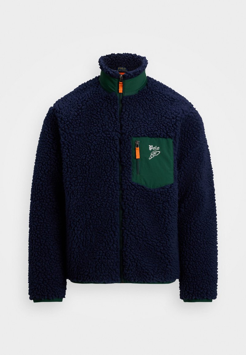 Veste en polaire bleu marine avec col haut, poche poitrine zippée verte et tirettes orange, conçue pour la chaleur et les activités de plein air.
