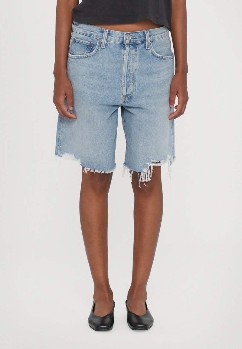 Shorts en denim bleu clair avec un ourlet effiloché, affichant une coupe décontractée et un design à cinq poches. Portés avec un top court noir et des ballerines noires.