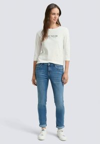 Γυναίκα με λευκή μακρυμάνικη μπλούζα με το λογότυπο "Tom Tailor", μπλε τζιν ρολέρ και λευκά αθλητικά παπούτσια, που στέκεται μπροστά σε ανοιχτό γκρι φόντο.