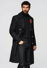 Schwarzer Wollmantel mit goldenen Beschlägen, doppelt geknöpftes Design, bestickter roter Fruchtakzent, getragen über einem schwarzen Rollkragenpullover und schwarzer Hose.