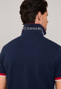Granatowa koszulka polo z wysokim kołnierzykiem, ozdobiona białymi i czerwonymi akcentami. Napis na kołnierzyku głosi "ACONNABL". Gładka bawełniana tkanina, luźny krój.