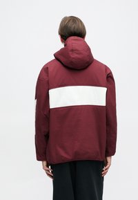 Casaco capuz bordeaux com uma risca horizontal branca nas costas. Feito de um tecido texturizado, apresenta um corte relaxado e design simples.