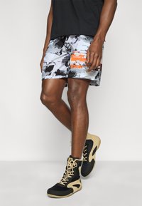 Ανδρικό μαγιό σορτς σε λευκό με μαύρα και πορτοκαλί αφηρημένα σχέδια, με ελαστική μέση και λεπτομέρεια με λογότυπο στο πλάι. Συνδυάζεται με μαύρα παπούτσια μποτάκια.