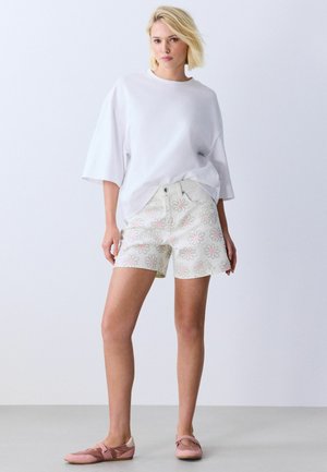 Blonde Frau, die ein übergroßes weißes T-Shirt, weiße Shorts mit pinken Blumenmotiven und pinke flache Schuhe trägt, steht auf einem grauen Boden.