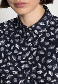 Camicia blu navy con un motivo a conchiglie bianche, colletto classico, bottoni sulla parte anteriore e una texture di tessuto liscia.