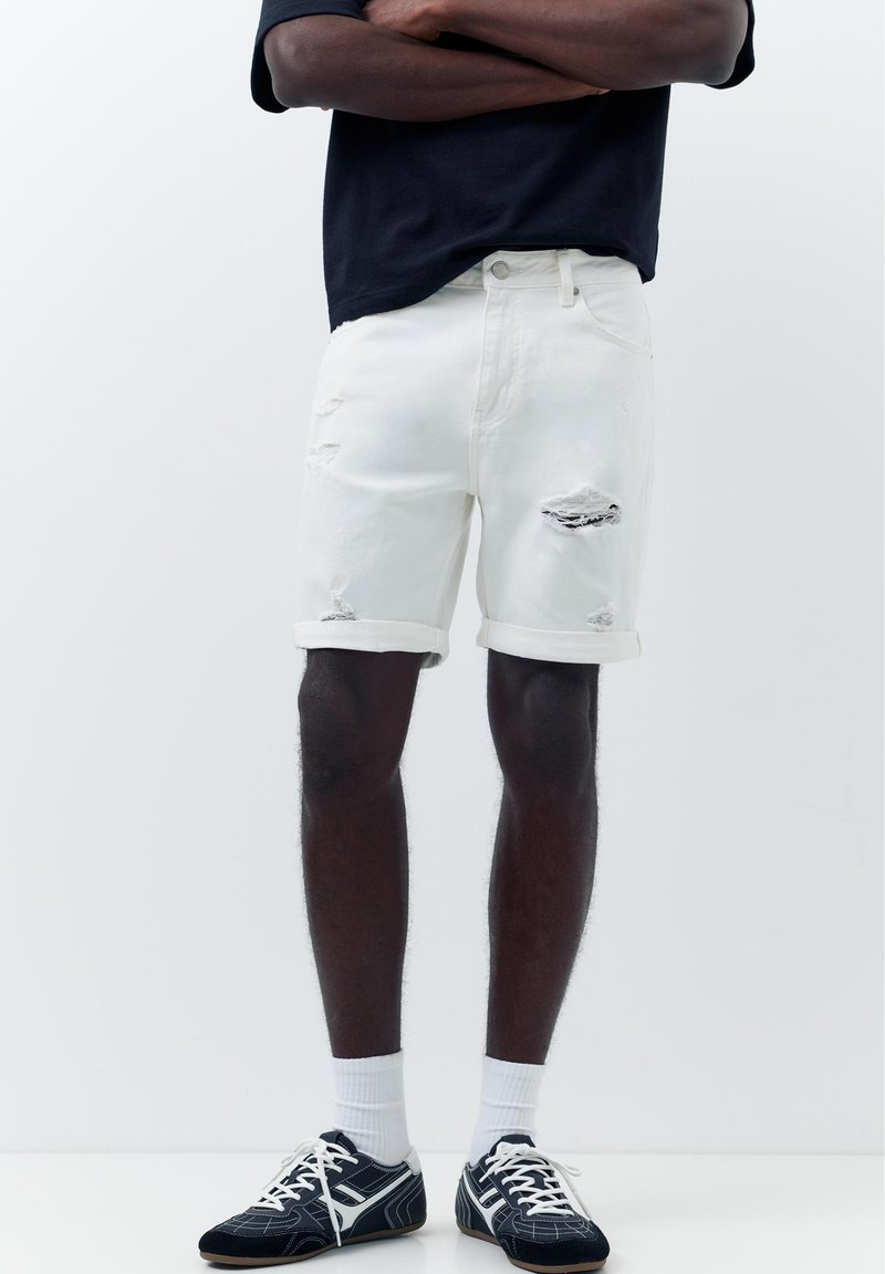 Personne debout, bras croisés, portant un short blanc déchiré, un t-shirt noir, des chaussettes blanches et des baskets noires et blanches sur un fond clair uni.