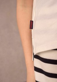 Bílá bavlněná tričko s červeným a námořnickým "Superdry" štítkem, spojené s černými a bílými pruhovanými kalhotami, které ukazují detaily švů a texturu materiálů.