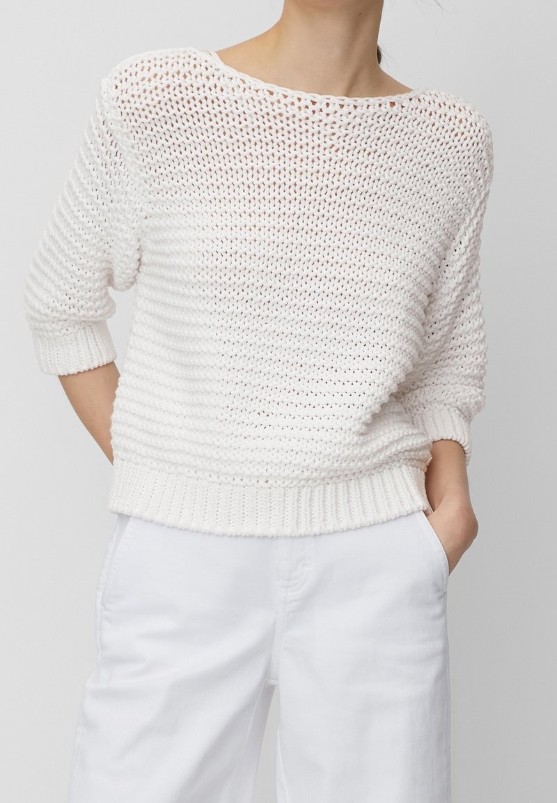 Femme portant un pull blanc en maille lâche à manches trois-quarts et un pantalon blanc, mains dans les poches, devant un fond uni.