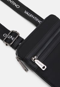 Geantă Valentino neagră cu textură de nailon, având două compartimente cu fermoar și curea reglabilă cu logo. Accente din metal argintiu.