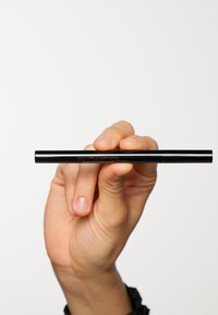 Penna per eyeliner nera tenuta tra le dita, con una finitura lucida e il marchio dorato "REVOLUTION" lungo il lato. Design cilindrico e liscio.
