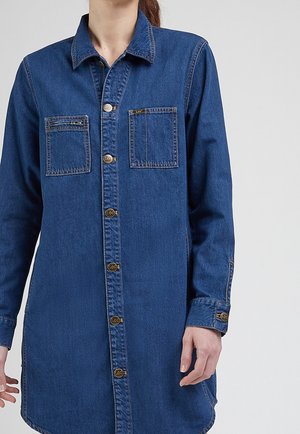 Kvinde iført langærmet blå denim-skjorte med knaplukning og to brystlommer, stående mod en ensfarvet lys baggrund.