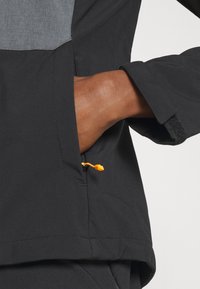 Veste noire avec un panneau supérieur gris, équipée d'une poche zippée et d'une languette de tirage orange. Le matériau est lisse, avec une texture ressentie sur la partie grise.