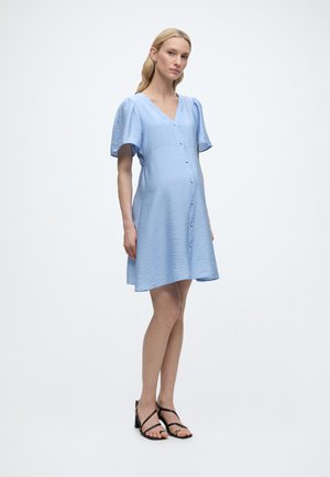 Rase naine hele sinise nööpidega kleidiga, lühikeste lendavate varrukatega, seisab mustade avatud varbaga kontsakingadega valgel taustal.