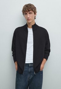 Schwarze Zip-Jacke mit hohem Kragen und gerippten Bündchen, kombiniert mit einem weißen T-Shirt und blauen Jeans. Glatte Stofftextur.