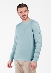 Felpa blu chiaro, a coste, con maniche lunghe e collo rotondo, con un piccolo tag nero sulla manica, abbinata a pantaloni beige.
