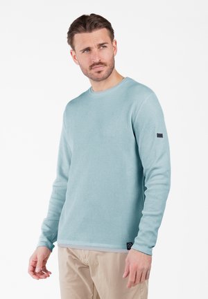 Sudadera de manga larga de color azul claro, con canalé, y cuello redondo, con una pequeña etiqueta negra en la manga, combinada con pantalones beige.