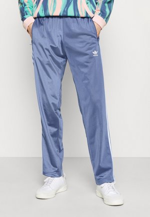 Person wearing blue Adidas track pants with white stripes and white sneakers, hands in pockets, against plain background.