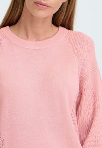Ljusrosa stickad tröja med rund hals och raglanärmar. Ärmarna har en mer öppen stickning som kontrasterar med blusens huvuddel.