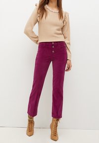 Pull en tricot beige avec des manches volantées, associé à un pantalon en daim violet avec trois boutons à l'avant. Bottes en cuir avec lacets couleur beige et accents dorés.