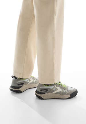 Zapatillas deportivas grises con suela moteada y cordones verde neón; combinadas con pantalones de pierna ancha color crema, que muestran un diseño casual y moderno.