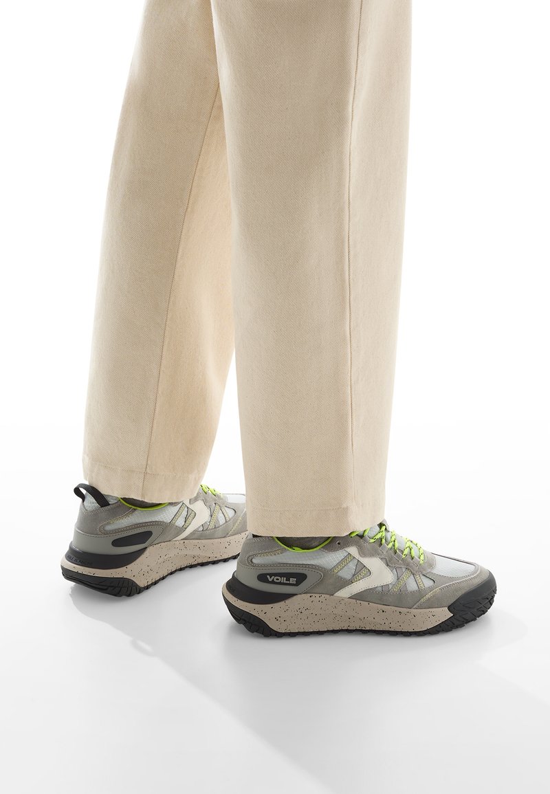 Scarpe da ginnastica grigie con suola chiazzata e lacci verde neon; abbinate a pantaloni a gamba larga color crema, che mostrano un design casual e moderno.