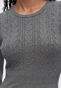Gros plan d'une personne portant un pull gris à torsades avec des motifs tressés verticaux et un col arrondi.