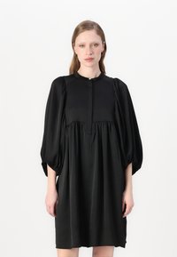 Vestido preto feito de tecido suave, com decote redondo, mangas bufantes e uma cintura franzida com um detalhe de botão discreto.