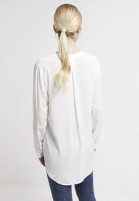 Langærmet hvid bluse med buet kant, lavet af letvægtsmateriale. Har et simpelt design med en lige ryg og knapmanchetter.