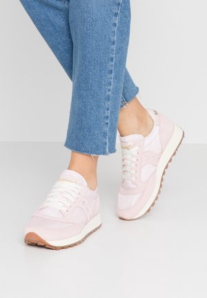 Persoon die lichtroze vetersneakers en een blauwe spijkerbroek draagt, staand op een glad wit oppervlak met één voet iets opgeheven.