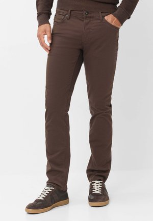 Mand iført brune slim-fit bukser, mørkebrun sweater og mørke sneakers med hvide snørebånd står foran en ensfarvet baggrund.